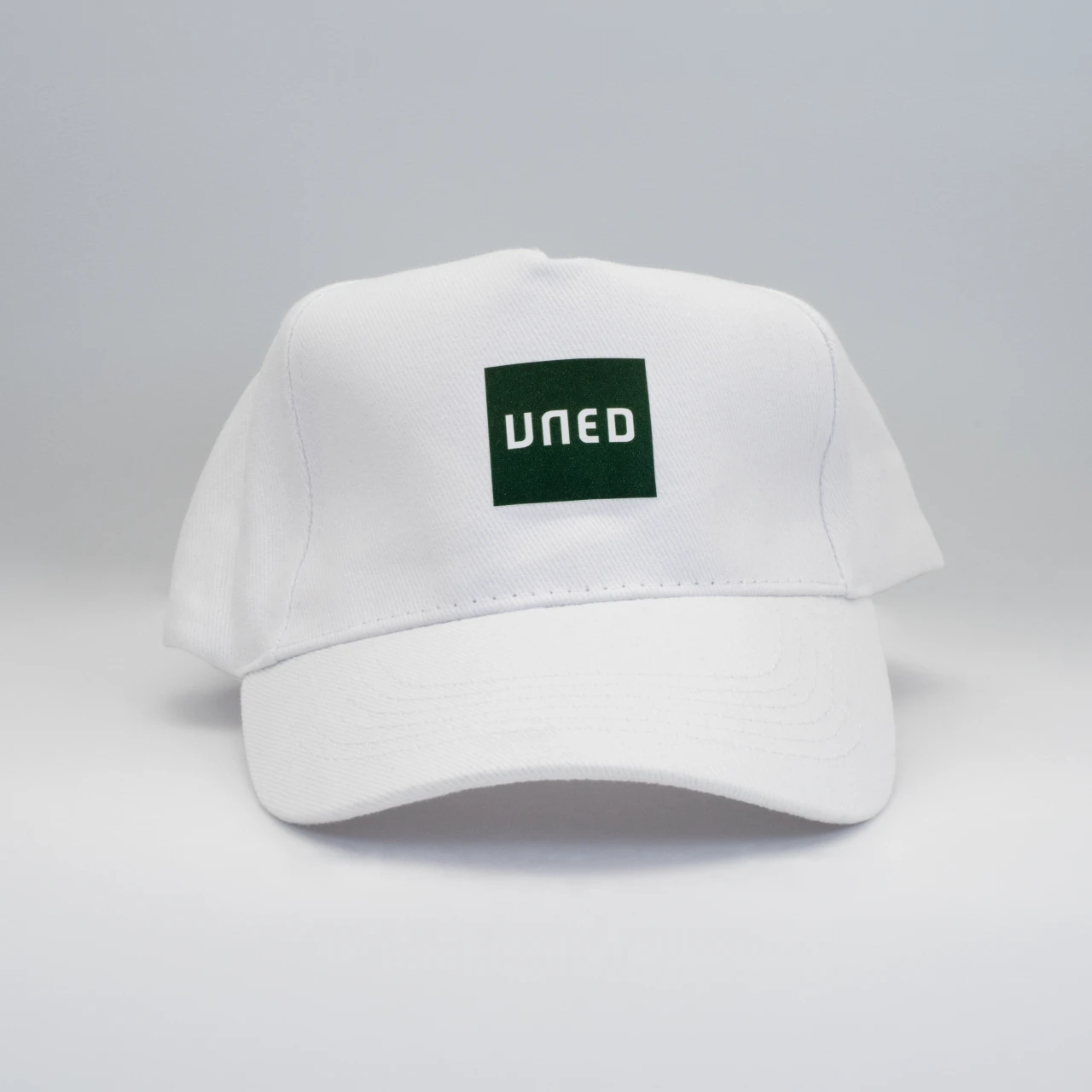 Gorra de cinco paneles en blanco con parche bordado UNED en verde, vista frontal sobre fondo gris.