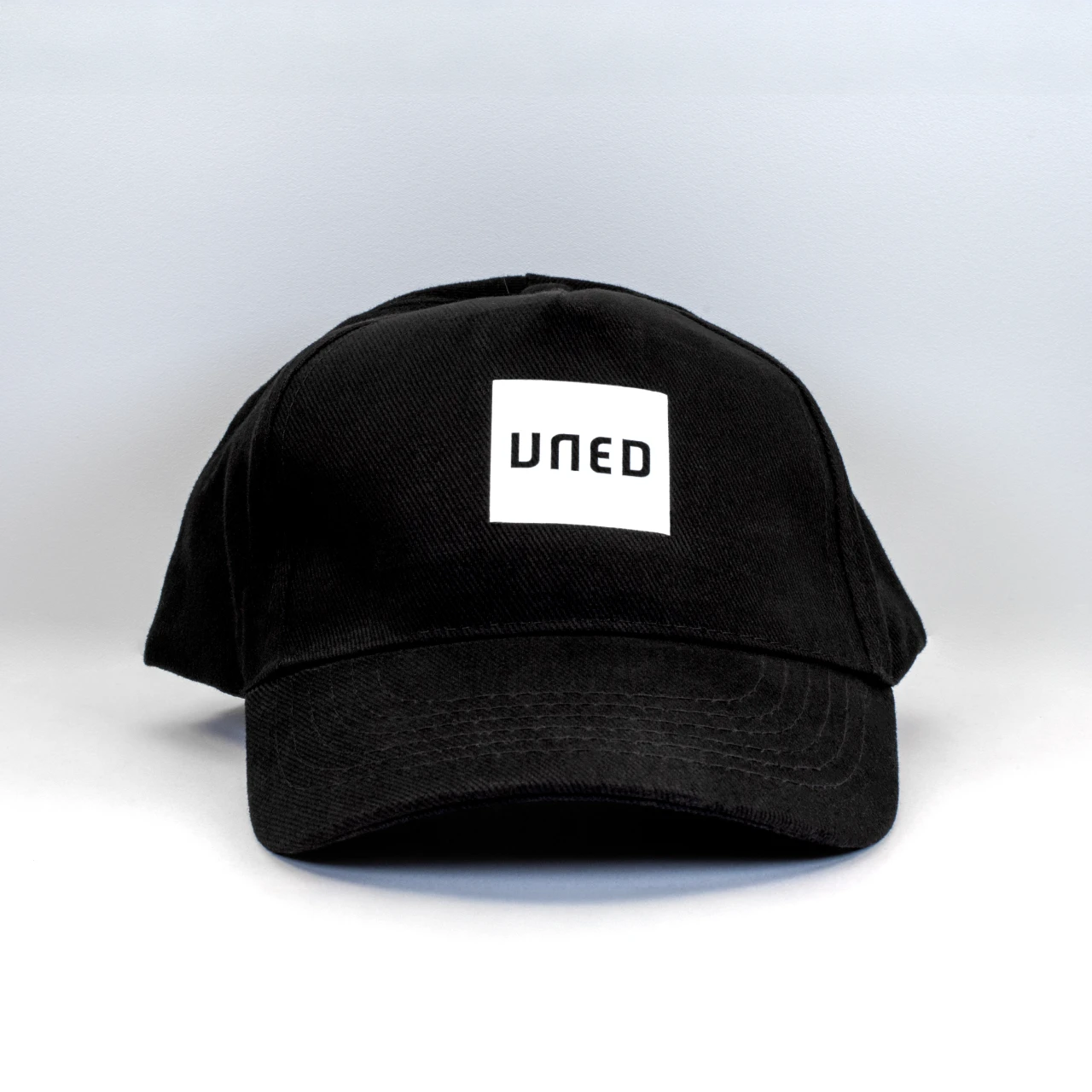 Gorra de cinco paneles en negro con parche bordado UNED, vista frontal sobre fondo gris. Gorra de cinco paneles en negro con parche bordado UNED, vista frontal sobre fondo gris.