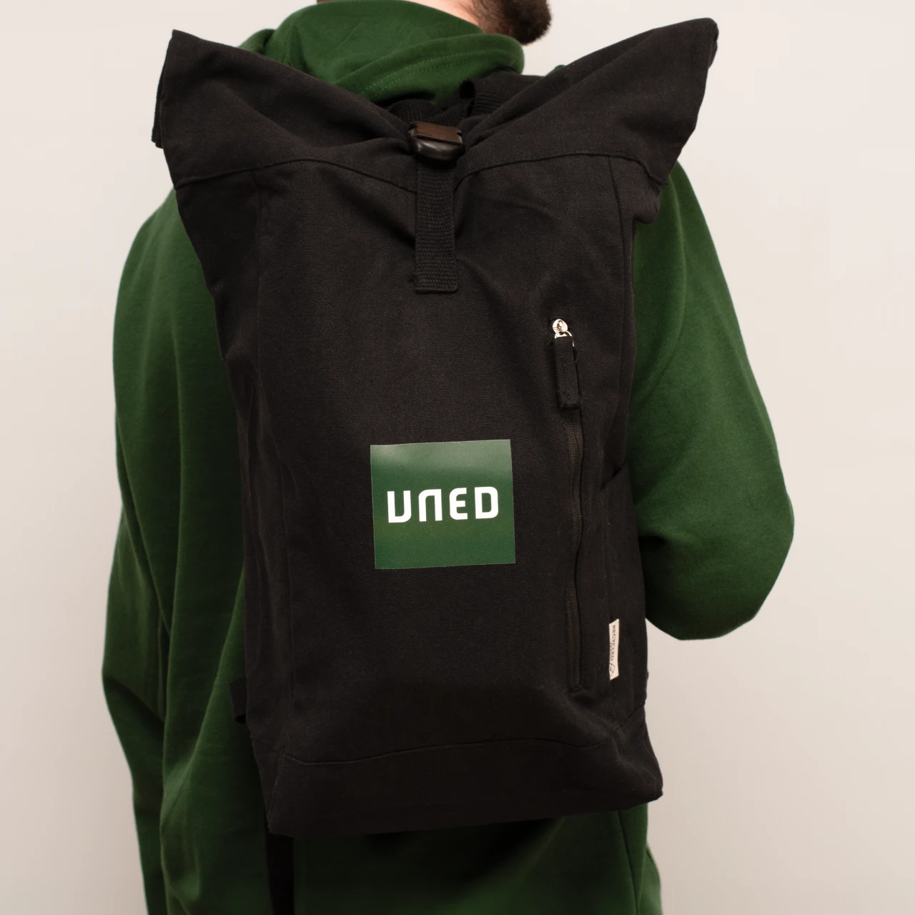 Detalle frontal de mochila roll-top en canvas reciclado negro con parche UNED en verde.