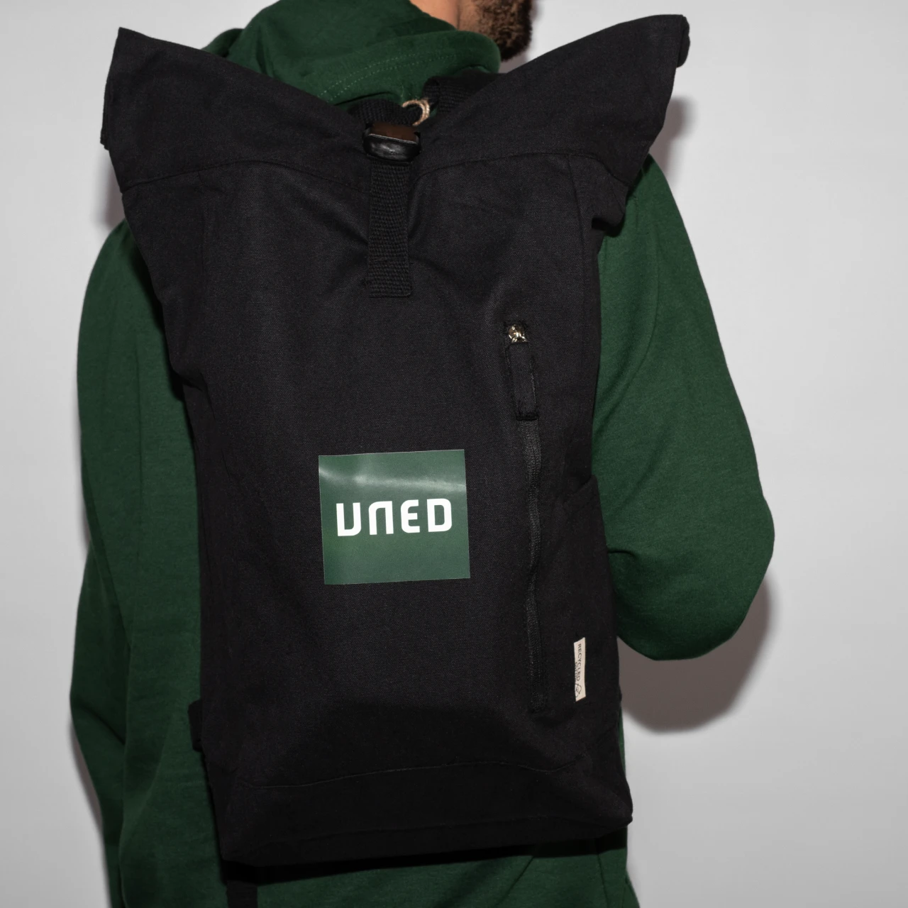 Detalle frontal de mochila roll-top en canvas reciclado negro con parche UNED en verde.