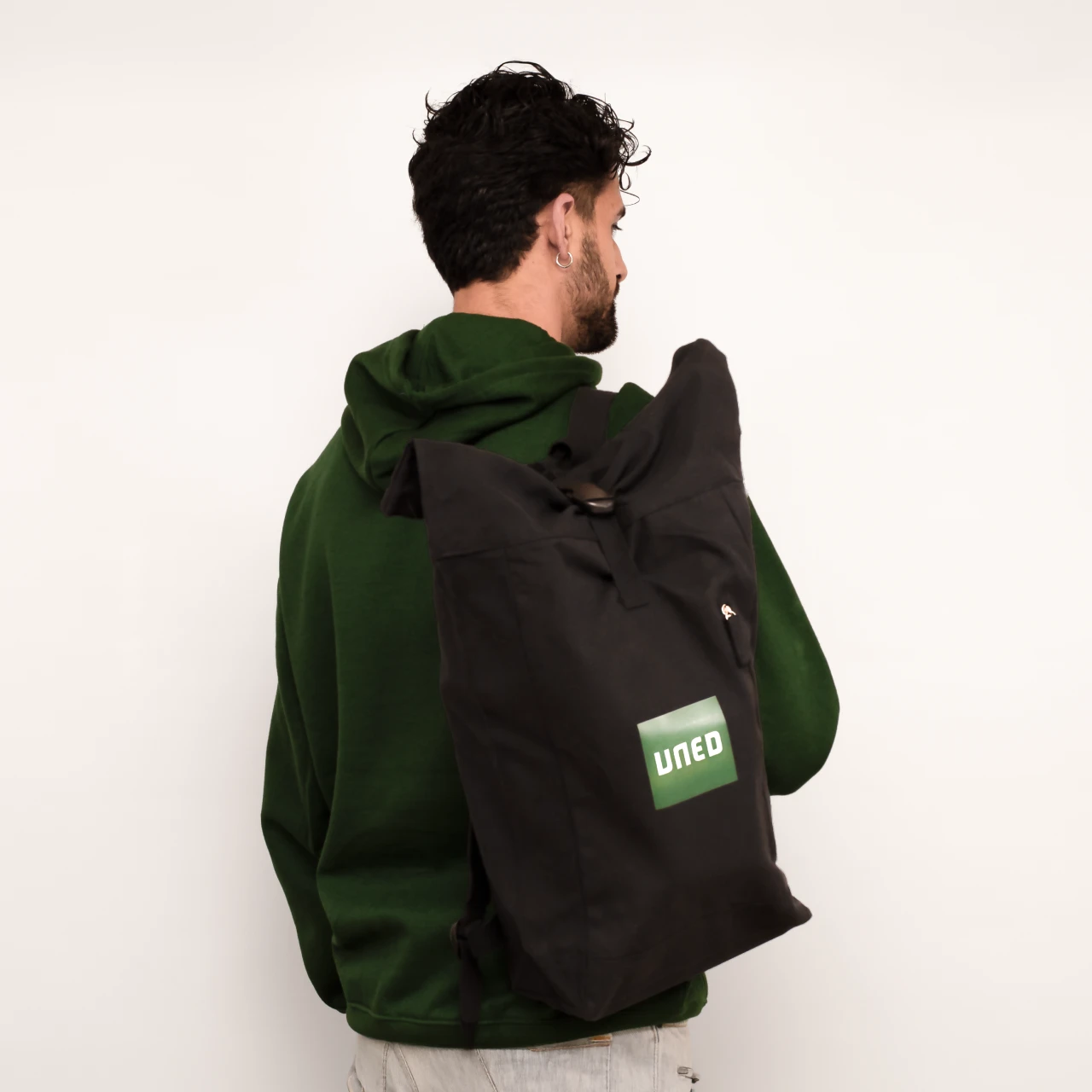 Mochila tipo roll-top en canvas reciclado negro con logo UNED en verde, vista de espaldas sobre modelo.