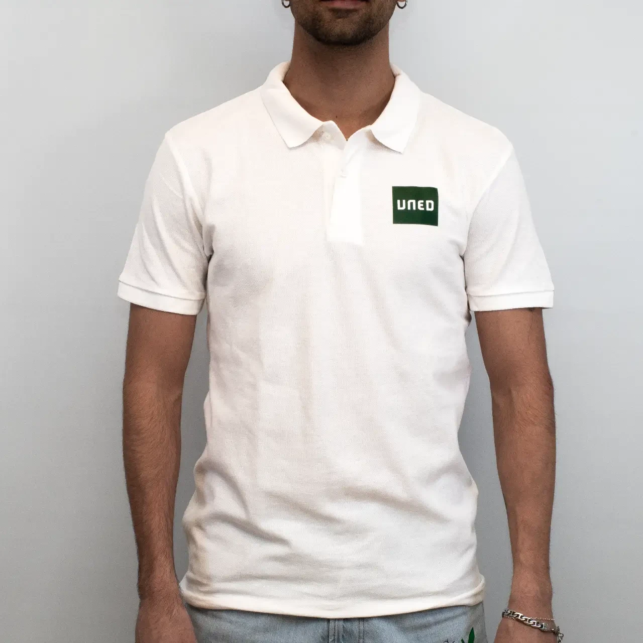 Polo de piqué blanco con parche UNED en verde, vista frontal sobre modelo.