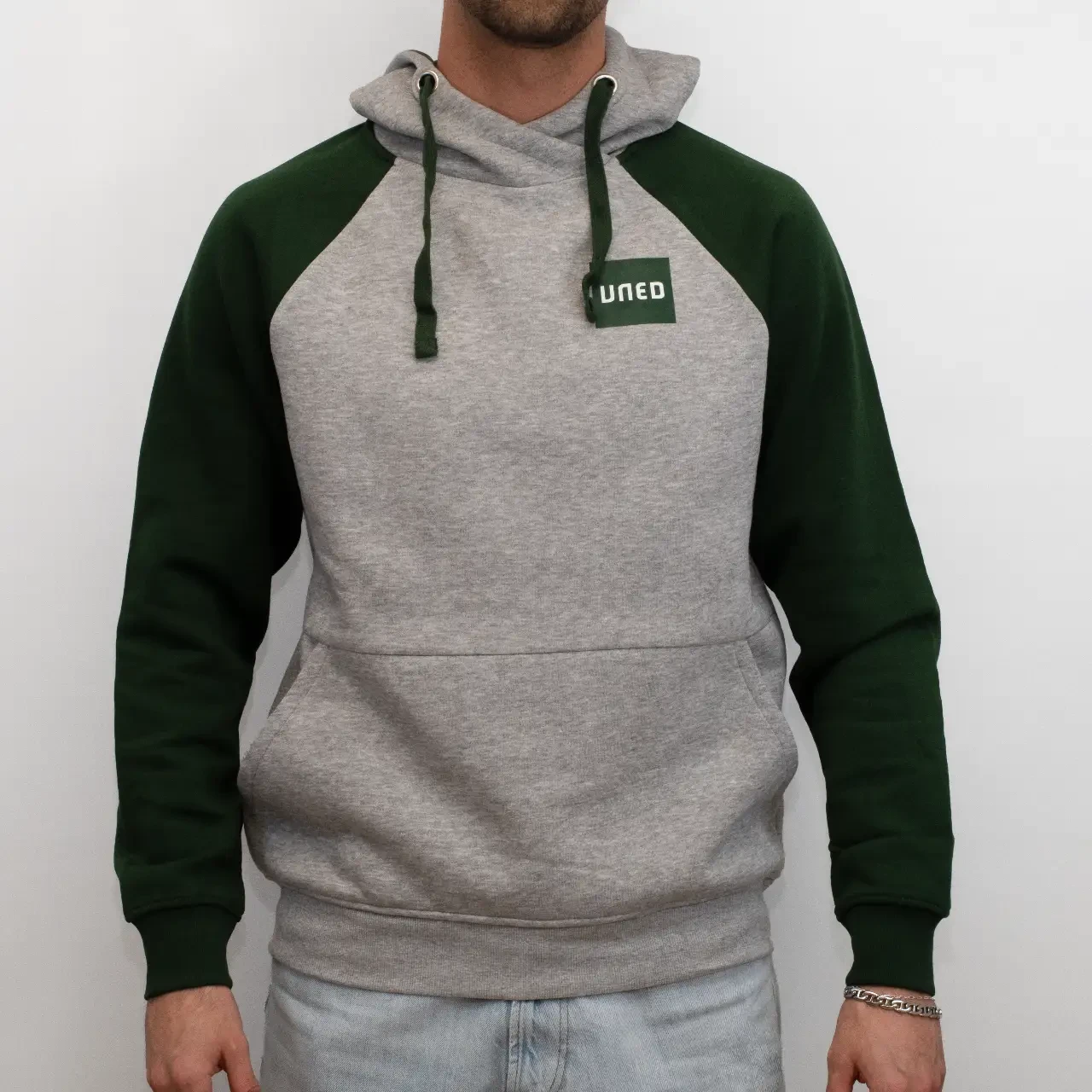 Sudadera bicolor con capucha en gris y mangas raglán en verde con parche UNED, vista frontal sobre modelo.