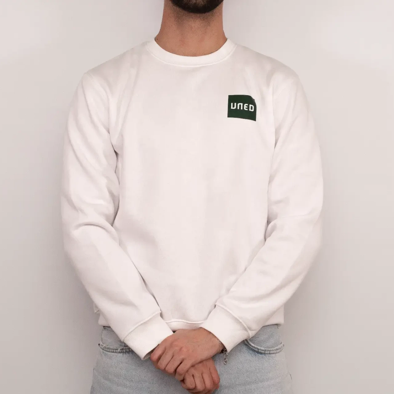 Sudadera unisex de cuello redondo en color blanco con parche UNED en verde, vista frontal sobre modelo.