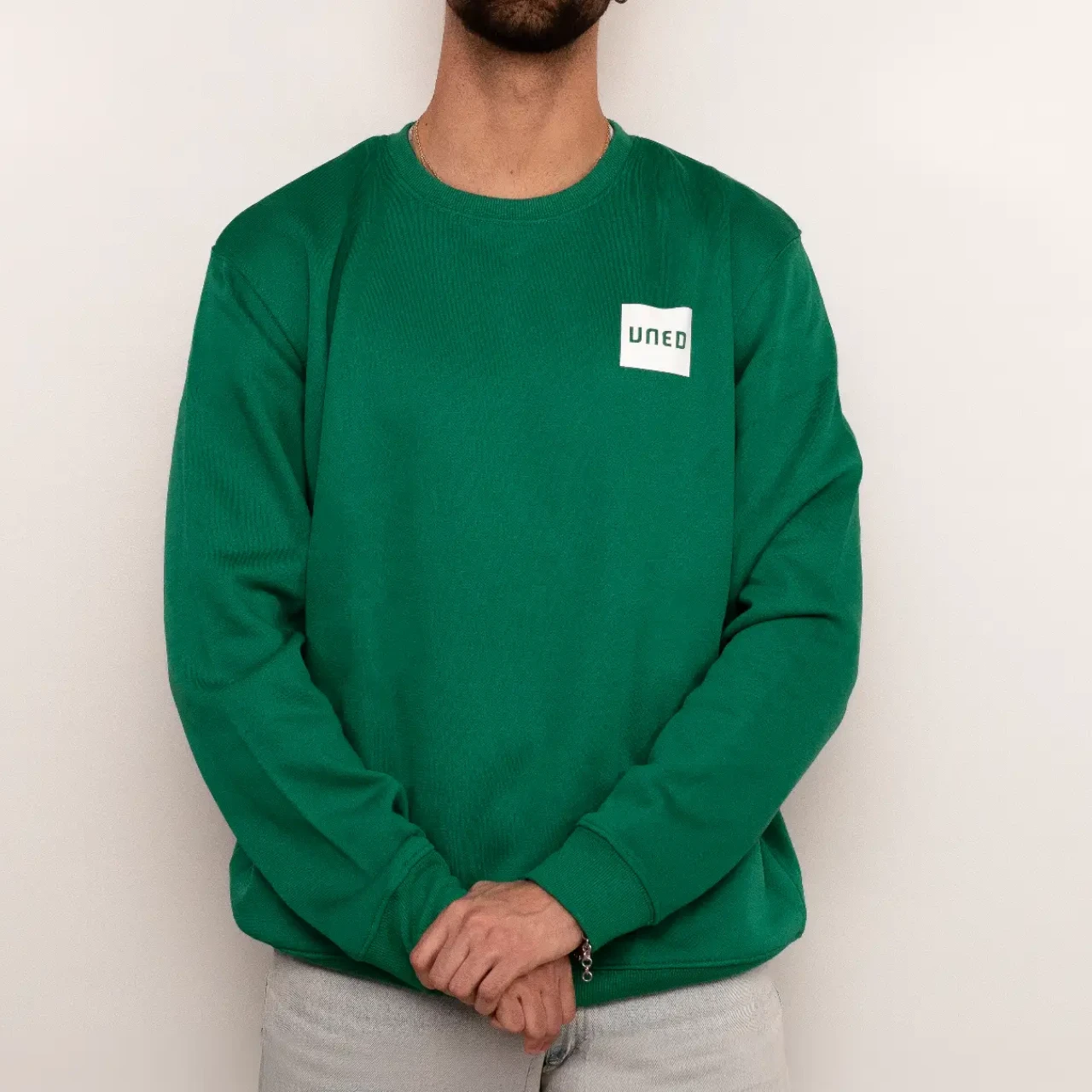 Sudadera unisex de cuello redondo en color verde con parche UNED en crudo, vista frontal completa sobre modelo.
